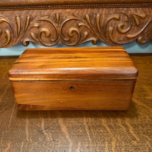May include: A rectangular wooden box with a hinged lid, crafted from light brown wood. The box features a small keyhole on the front and is set against a backdrop of ornate, carved wooden details. The box is likely used for storage.