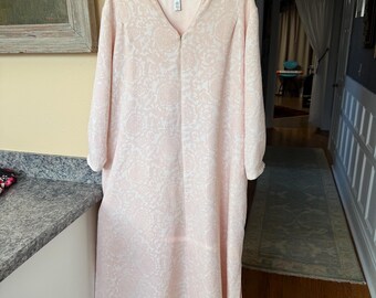 May include: A light pink, long-sleeved dress with a V-neck and a white floral pattern. The dress is hanging on a white hanger. The dress is made of a soft, flowing fabric.
