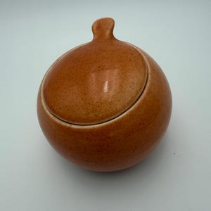 May include: A spherical ceramic sugar bowl with a speckled terracotta orange glaze. The container features a matching rounded lid with a small, upright handle at the top, creating a smooth, uniform silhouette with a glossy, textured finish.