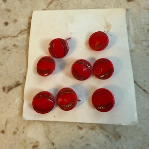 May include: Nine red buttons with a decorative design, arranged on a piece of off-white paper. The buttons have a glossy finish and a detailed, textured pattern in a contrasting color. The buttons are round and appear to be vintage.