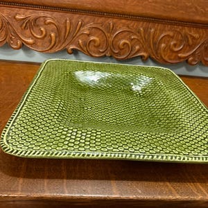 May include: A square, green ceramic serving dish with a textured, honeycomb-like pattern. The dish has a slightly raised center and a decorative border. The dish is displayed on a dark wooden surface.