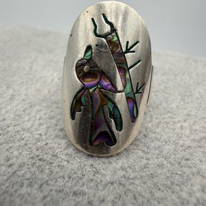 Könnte beinhalten: Ovale Silberring mit einem Vogeldesign und schillerndem Abalone-Muschel-Inlay. Der Vogel sitzt auf einem Ast, mit ausgeschnittenen Details. Der Ring hat eine polierte Oberfläche und eine Vintage-Ästhetik.