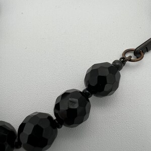 May include: Close-up of a black beaded necklace. The beads are faceted, reflecting light, and are connected by a dark metal clasp. The necklace is set against a white background.