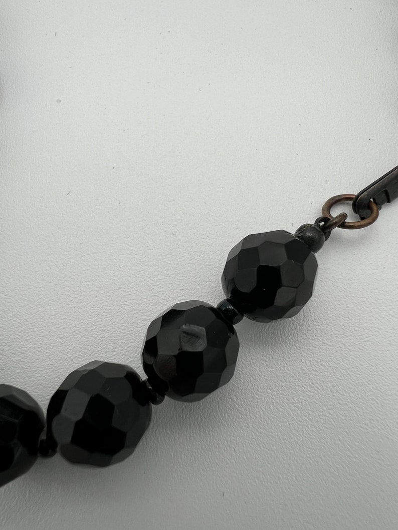 May include: Close-up of a black beaded necklace. The beads are faceted, reflecting light, and are connected by a dark metal clasp. The necklace is set against a white background.