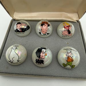May include: A set of six vintage cartoon character collectible marbles in a gray box. Each iridescent marble features a different character illustration, including Fosdick, Li'l Abner, and Pappy Yokum. The marbles are from Capp Ent., Inc.