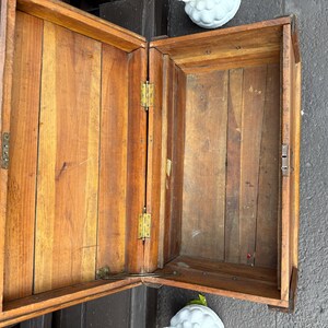 Puede incluir: Un cofre de madera abierto con tapa con bisagras, que muestra una c&aacute;lida veta de madera natural. El cofre presenta herrajes de lat&oacute;n, incluidas bisagras y un asa. El interior tambi&eacute;n es de madera, con tablones verticales visibles.