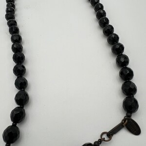 May include: A black beaded necklace with faceted beads. The necklace has a clasp and a small tag. The beads are a deep black color and have a shiny finish. The necklace is displayed against a white background.