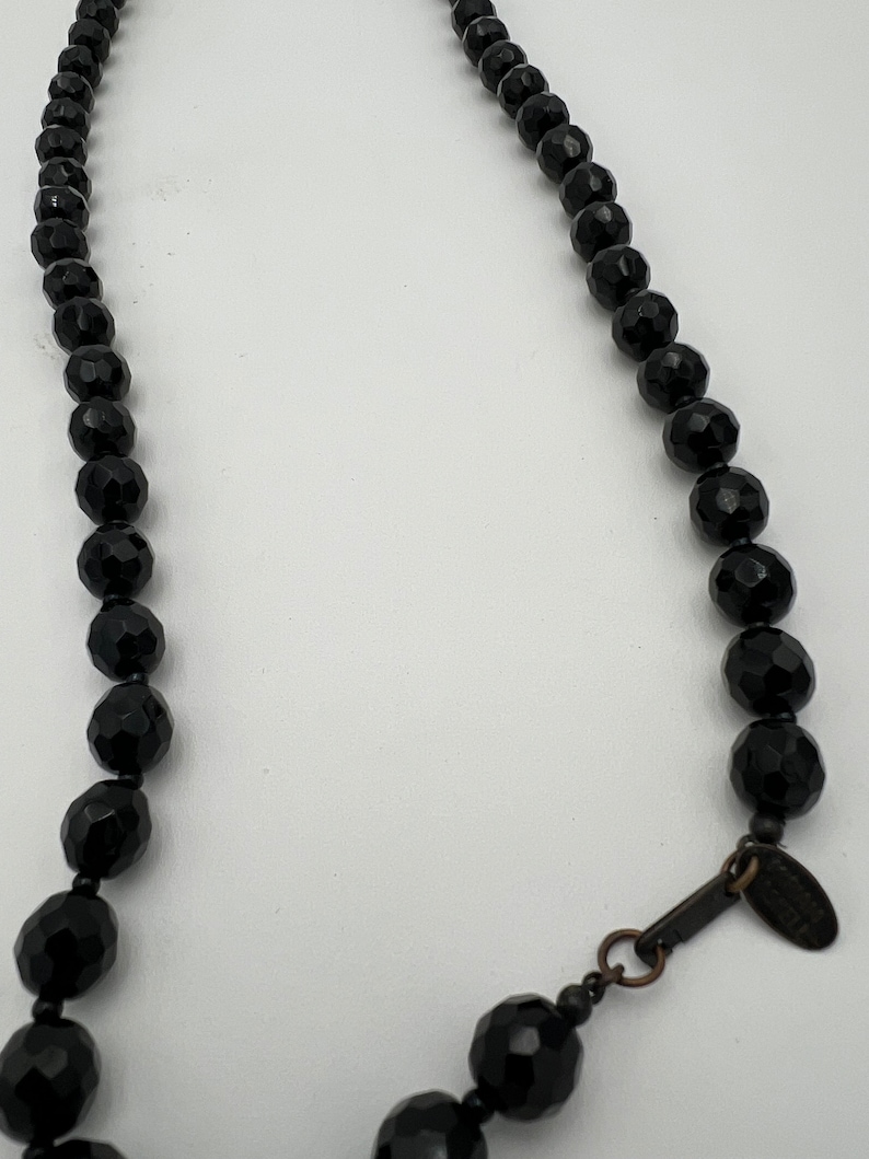 May include: A black beaded necklace with faceted beads. The necklace has a clasp and a small tag. The beads are a deep black color and have a shiny finish. The necklace is displayed against a white background.