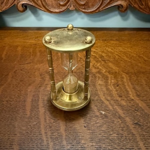 May include: A brass hourglass featuring a clear glass bulb filled with light-colored sand. The frame consists of a circular top and base connected by three slender, turned pillars. The top plate is secured with three small, rounded brass finials.