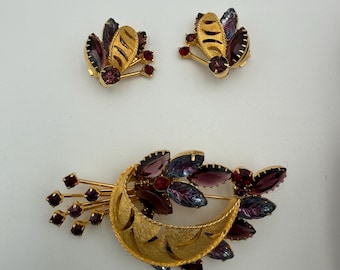 Peut inclure: Un ensemble de bijoux vintage comprenant une broche dorée et des boucles d'oreilles assorties. La broche a un motif de croissant avec des strass violets et rouges. Les boucles d'oreilles et la broche ont un motif de feuilles et de baies.