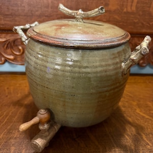 May include: A handmade ceramic water dispenser with a lid and a spigot. The stoneware pot is a muted green with a brown rim and lid. The handles and lid handle are designed to look like branches.