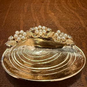 May include: A gold-colored, shell-shaped trinket dish. The dish has a textured surface with a scalloped design. The top edge is adorned with clusters of faux pearls and clear rhinestones. The dish is likely made of metal and is suitable for holding small items.