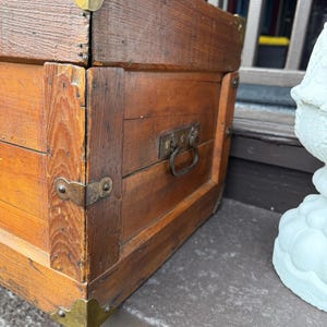 Puede incluir: Un ba&uacute;l de madera vintage con protectores de esquina de lat&oacute;n y un asa de metal. El ba&uacute;l es de color marr&oacute;n c&aacute;lido y parece desgastado, lo que sugiere edad y uso. El ba&uacute;l probablemente se utiliza para el almacenamiento.