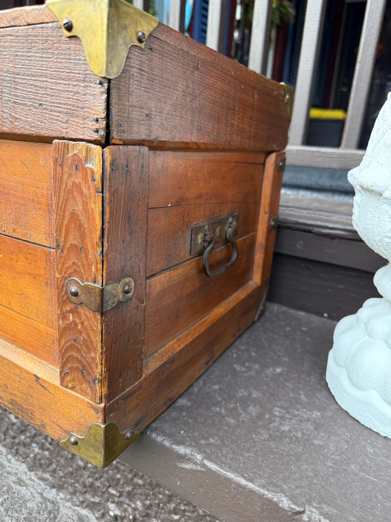 Puede incluir: Un ba&uacute;l de madera vintage con protectores de esquina de lat&oacute;n y un asa de metal. El ba&uacute;l es de color marr&oacute;n c&aacute;lido y parece desgastado, lo que sugiere edad y uso. El ba&uacute;l probablemente se utiliza para el almacenamiento.