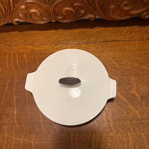 May include: A white, round lid with a dark brown handle. The lid has two small handles on either side. The lid is sitting on a wooden surface.
