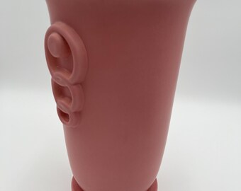 May include: A dusty rose-colored vase with a flared top and a circular base. The vase features a decorative, raised swirl design on one side. The vase is made of a matte material and stands on a round base.