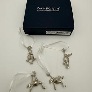 May include: A set of four silver-colored metal ornaments shaped like acrobatic figures, each suspended by a white ribbon. The ornaments are presented with a navy blue box that has the text "DANFORTH we work metal by hand".
