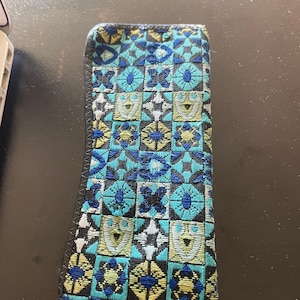 May include: A rectangular case with a geometric pattern in shades of blue, yellow, and black. The case has a stitched black border. The pattern is made up of squares and floral designs.