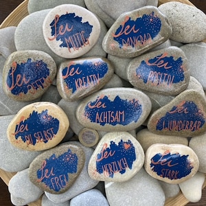 Könnte beinhalten: Eine Sammlung bemalter Steine mit dem Wort "Sei" (deutsch für "sei") und verschiedenen positiven Affirmationen auf Deutsch, wie "mutig", "dankbar" und "kreativ". Die Steine sind in Blautönen, Weiß und Rosa bemalt.