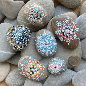 Puede incluir: Seis piedras pintadas con diseños de mandalas. Las piedras son de varios tonos de marrón y presentan patrones de puntos coloridos en azul, verde, rosa, naranja y amarillo.