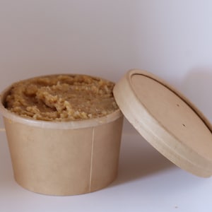 May include: A brown paper container filled with a light brown, creamy substance, with a matching lid off to the side.