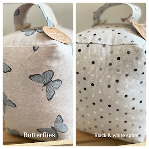 May include: Two doorstop cushions, one with a butterfly pattern and the other with black and white polka dots. Both cushions are made of a light beige fabric and have a loop handle.