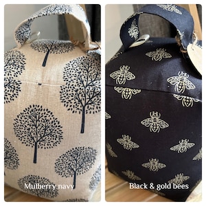 May include: Two doorstop cushions. One is beige with a black tree pattern and the text "Mulberry-navy". The other is black with a gold bee pattern and the text "Black & gold bees".