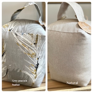 May include: Two doorstop cushions, one with a grey and white peacock feather pattern and the other in a natural beige fabric. Both have a loop handle.