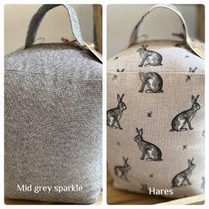 May include: Two doorstop cushions. One is a solid mid-grey color with a sparkle finish. The other is a beige fabric with a repeating pattern of black hares. The text "Hares" is printed below the cushion.