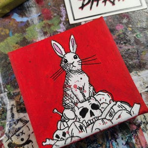 Op de afbeelding: Een vierkant canvas schilderij met een wit konijn dat op een stapel schedels zit tegen een felrode achtergrond. Het kunstwerk gebruikt zwarte inktcontouren en rode verfspatten. Een witte kaart met de tekst SOMETHING DIFRNT is zichtbaar.