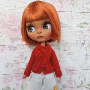 Puede incluir: Un c&aacute;rdigan de punto rojo con botones dorados, usado por una mu&ntilde;eca con cabello naranja y ojos azules. La mu&ntilde;eca lleva pantalones blancos con parches.