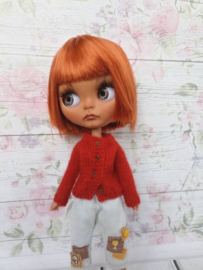Puede incluir: Un c&aacute;rdigan de punto rojo con botones dorados, usado por una mu&ntilde;eca con cabello naranja y ojos azules. La mu&ntilde;eca lleva pantalones blancos con parches.