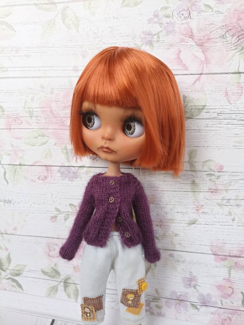 Puede incluir: Una mu&ntilde;eca con el pelo naranja que lleva un c&aacute;rdigan de punto morado y pantalones azul claro con parches.