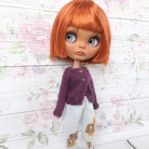 Puede incluir: Una mu&ntilde;eca con el pelo naranja que lleva un c&aacute;rdigan de punto morado y pantalones azul claro con parches marrones. La mu&ntilde;eca tiene ojos azules y una expresi&oacute;n neutral.