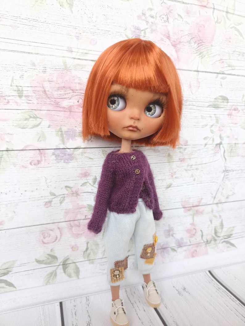Puede incluir: Una mu&ntilde;eca con el pelo naranja que lleva un c&aacute;rdigan de punto morado y pantalones azul claro con parches marrones. La mu&ntilde;eca tiene ojos azules y una expresi&oacute;n neutral.