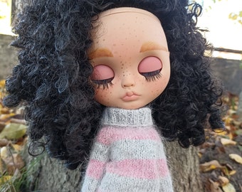 May include: A doll with long, curly black hair wearing a grey and pink striped sweater and blue jeans. The doll has closed eyes and freckles on its face.