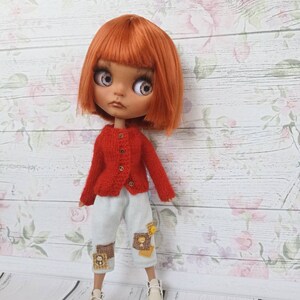 Puede incluir: Una mu&ntilde;eca con el pelo rojo que lleva un c&aacute;rdigan de punto rojo, pantalones azules claros parchados y zapatos blancos. La mu&ntilde;eca est&aacute; de pie sobre un fondo de madera blanco con un patr&oacute;n floral.
