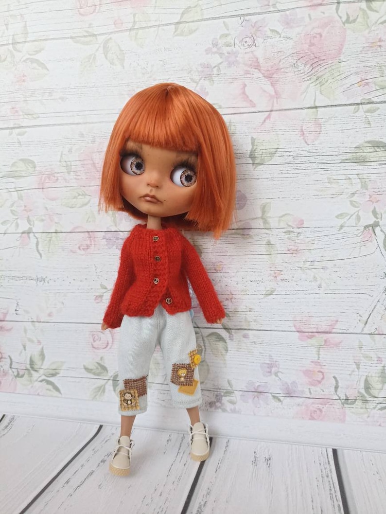 Puede incluir: Una mu&ntilde;eca con el pelo rojo que lleva un c&aacute;rdigan de punto rojo, pantalones azules claros parchados y zapatos blancos. La mu&ntilde;eca est&aacute; de pie sobre un fondo de madera blanco con un patr&oacute;n floral.
