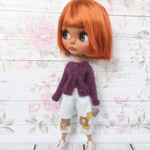 Puede incluir: Una mu&ntilde;eca con cabello naranja que lleva un c&aacute;rdigan morado, pantalones vaqueros azules claros con parches y zapatillas blancas. La mu&ntilde;eca est&aacute; de pie sobre una superficie de madera blanca con un fondo de patr&oacute;n floral.