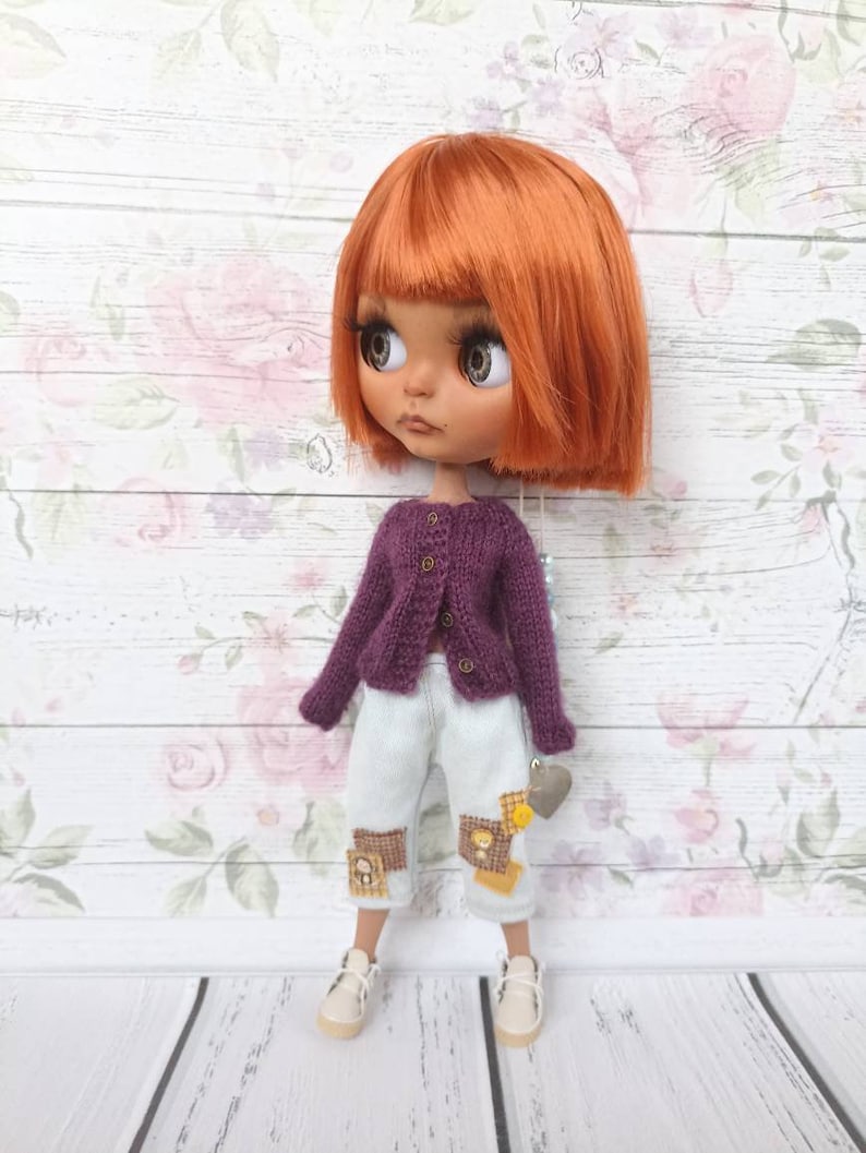 Puede incluir: Una mu&ntilde;eca con cabello naranja que lleva un c&aacute;rdigan morado, pantalones vaqueros azules claros con parches y zapatillas blancas. La mu&ntilde;eca est&aacute; de pie sobre una superficie de madera blanca con un fondo de patr&oacute;n floral.