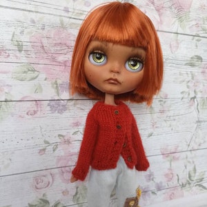 Puede incluir: Una mu&ntilde;eca con el pelo rojo que lleva un c&aacute;rdigan de punto rojo y pantalones azules claros con parches. La mu&ntilde;eca tiene ojos verdes y una expresi&oacute;n neutral.