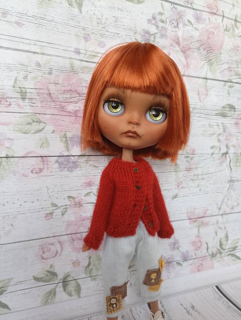 Puede incluir: Una mu&ntilde;eca con el pelo rojo que lleva un c&aacute;rdigan de punto rojo y pantalones azules claros con parches. La mu&ntilde;eca tiene ojos verdes y una expresi&oacute;n neutral.