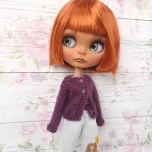Puede incluir: Una mu&ntilde;eca con cabello naranja que lleva un c&aacute;rdigan de punto morado y pantalones azul claro con parches bordados.