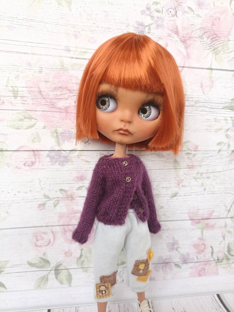 Puede incluir: Una mu&ntilde;eca con cabello naranja que lleva un c&aacute;rdigan de punto morado y pantalones azul claro con parches bordados.