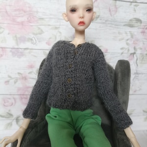 May include: A doll wearing a gray knitted cardigan with three brown buttons and green pants. The doll has a pale complexion, red lips, and dark eye makeup. The doll is seated in a gray chair.