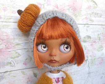 May include: A doll with orange hair wearing a gray knitted hat with a brown knitted pumpkin on top. The doll is wearing a white shirt with a black and white graphic print, a yellow cardigan, and white pants.