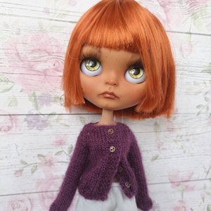 Puede incluir: Una mu&ntilde;eca con el pelo naranja que lleva un c&aacute;rdigan de punto morado con botones dorados y pantalones blancos.
