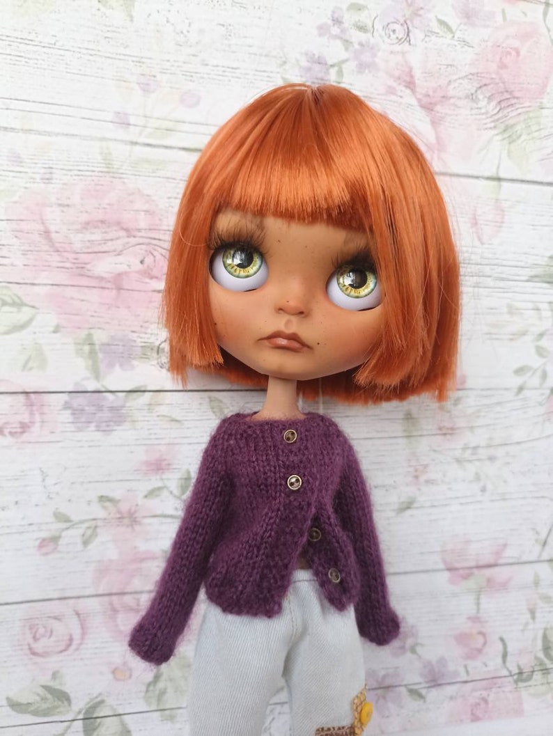 Puede incluir: Una mu&ntilde;eca con el pelo naranja que lleva un c&aacute;rdigan de punto morado con botones dorados y pantalones blancos.