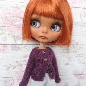 Puede incluir: Una mu&ntilde;eca con el pelo corto y de color naranja brillante que lleva un c&aacute;rdigan de punto morado con botones dorados y pantalones blancos.