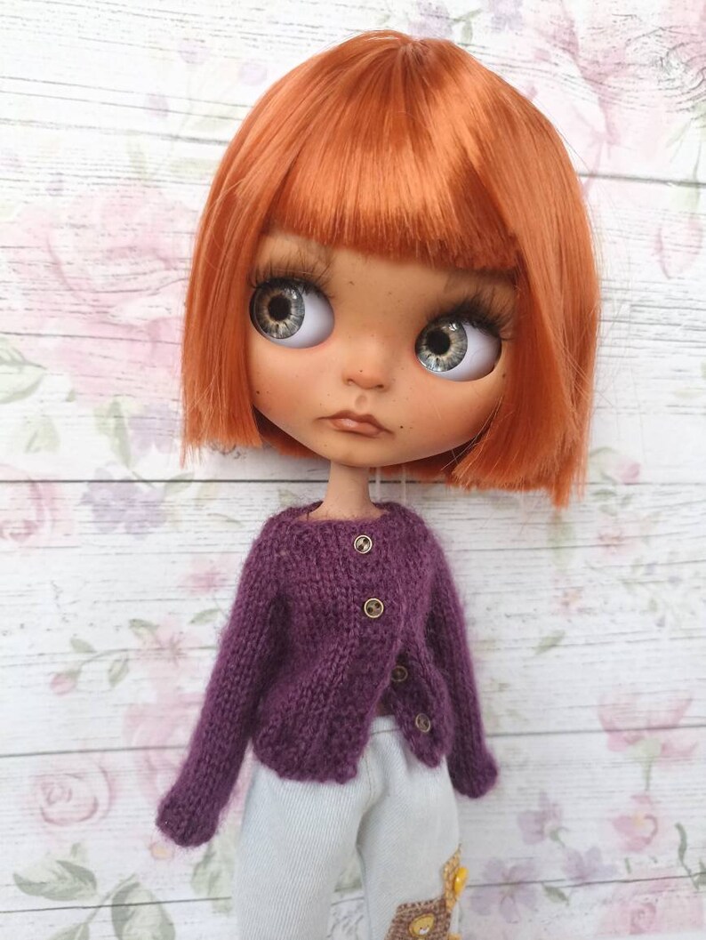 Puede incluir: Una mu&ntilde;eca con el pelo corto y de color naranja brillante que lleva un c&aacute;rdigan de punto morado con botones dorados y pantalones blancos.
